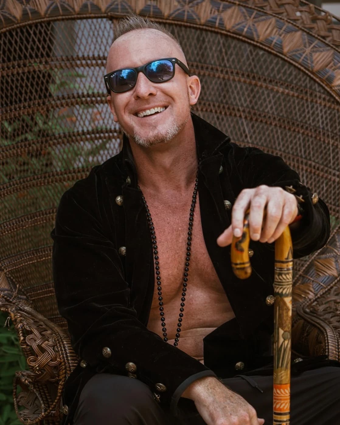 Matthew Layman seated in a peacock chair holding a carved didgeridoo.